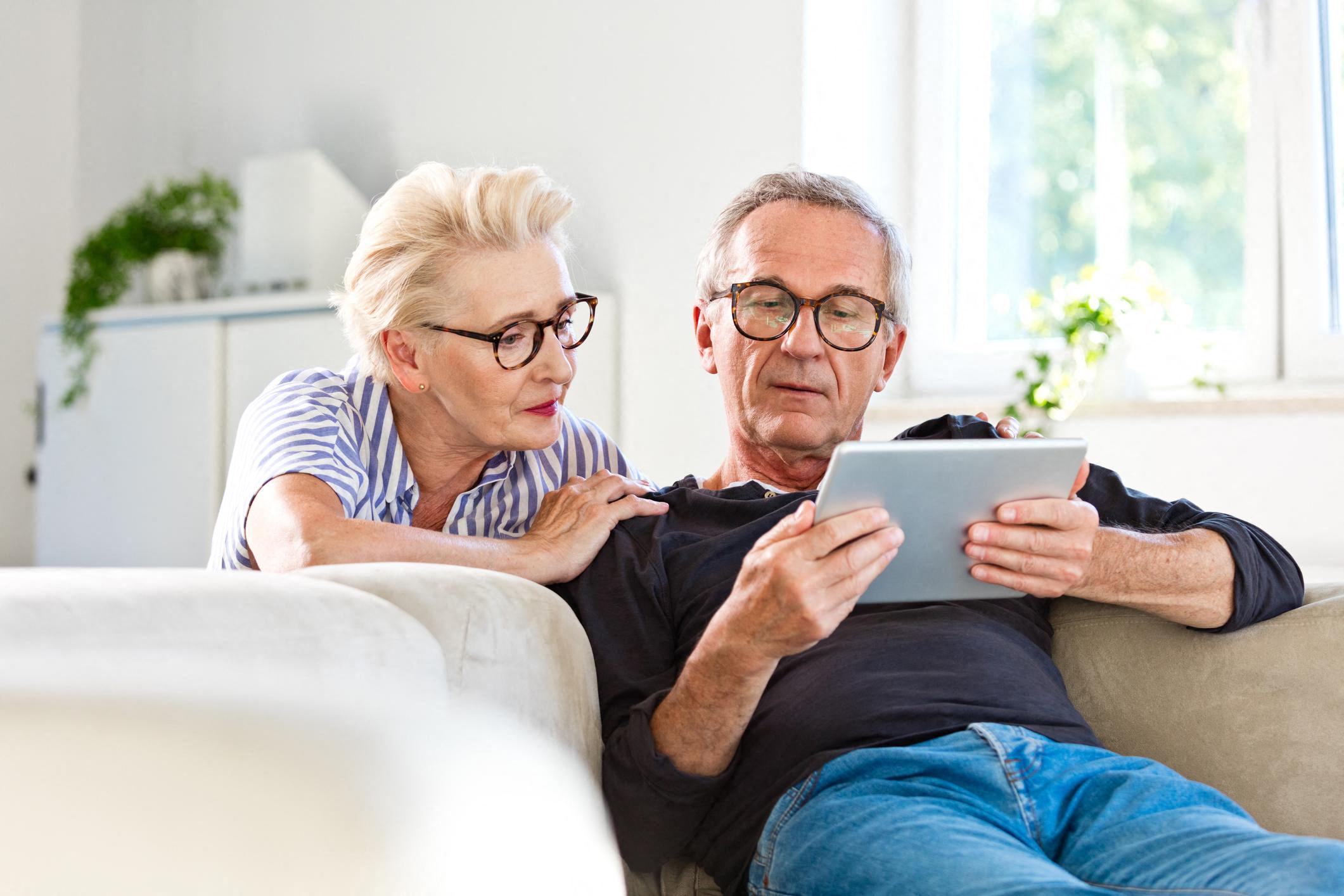 an older man and woman sitting on a couch looking at a tablet